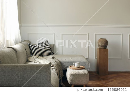 Elegant living room decor. Modern Scandinavian interior design. Cup of tea, coffee on tray, foot stool. Grey sofa, wooden pedestal with textured vase. Beautiful classic wall molding. Empty copy space. 126884931