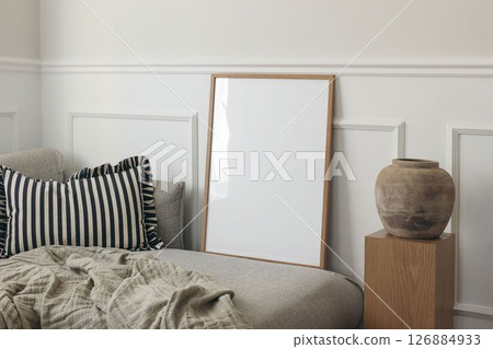 Blank thin wooden picture frame leaning against white stucco molding wall. Minimal elegant poster mockup. Artistic template. Grey sofa, ruffled pillow. Wooden pedestal, clay vase. Scandinavian 126884933