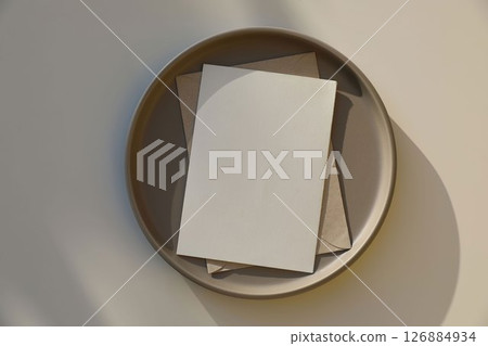 Elegant stationery composition. vertical blank greeting card, invitation, mock up scene. Brown ceramic plate. Beige table background in sunlight, shadows, flat lay, top view. Minimal still life. Elegant stationery composition. vertical blank greeting card, invitation, mock up scene. Brown ceramic plate. Beige table background in sunlight, shadows, flat lay, top view. Minimal still life. 126884934