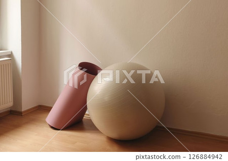 Closeup of exercise ball and pink yoga mat in soft ligh. Gym setting. Beige wall background, wooden floor. Fitness and relaxation concept. Training in studio. Set of sports equipment. Closeup of exercise ball and pink yoga mat in soft ligh. Gym setting. Beige wall background, wooden floor. Fitness and relaxation concept. Training in studio. Set of sports equipment. 126884942