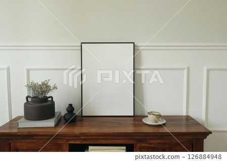 Minimalist home interior. Blank black picture frame mockup. Rustic wooden sideboard, dried flowers in a vase, and ceramic coffee cup. Neutral tones, natural light. Elegant still life. Poster display. 126884948