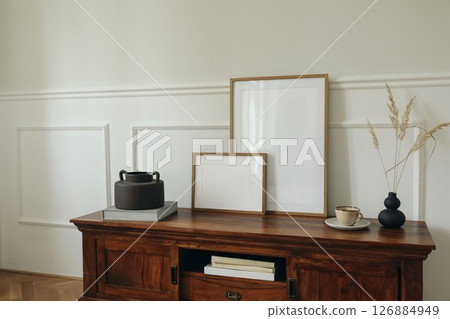 Scandinavian interior design. Modern living room. Set of blank picture mock up poster frames. Mahogany wooden dresser sideboard. Black vases with dry grass. Cup of tea, coffee. White wall background. 126884949