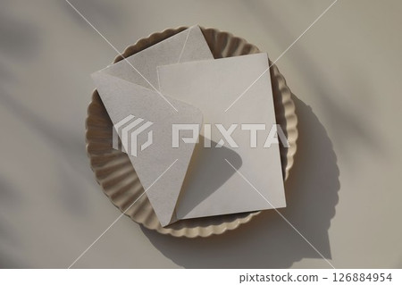 Beige fluted ceramic plate. Blank card, invitation mockup. Envelope in natural paper, softly lit by sunlight, shadows. Branding, birthday, wedding stationery. Elegant flat lay, neutral background. 126884954