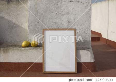 Blank wooden picture frame leaning against textured wall on Spanish house rooftop. Two yellow lemons on a concrete ledge. Natural sunlight, rustic setting, calm mood. Minimalist summer outdoor scene. 126884955