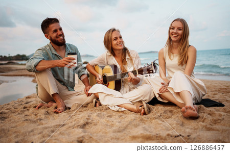 A group of friends joyfully enjoying music together while relaxing on a beach during sunset 126886457