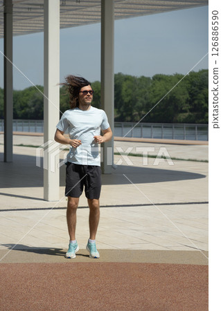Fit handsome caucasian brunette man wearing sports casual clothes doing warm up exercise and jogging on outdoor public park. Summer activity. Healthy lifestyle. 126886590