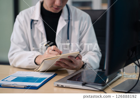 Male medicine doctor , physician or practitioner in lab room writing on blank notebook and work on laptop computer with medical stethoscope on the desk at hospital. Medic tech concept. 126888192