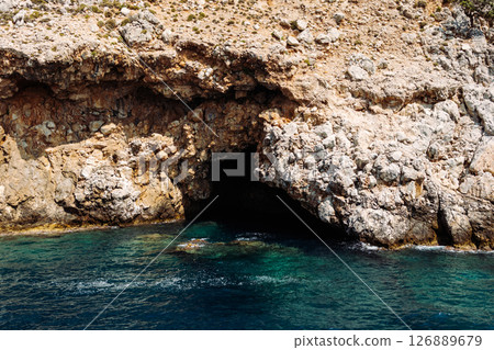 Sea cave in a rocky shore with turquoise transparent water. Picturesque natural landscape with rough texture of stones and deep entrance to the grotto. Ecotourism. Fascinating seascape. 126889679