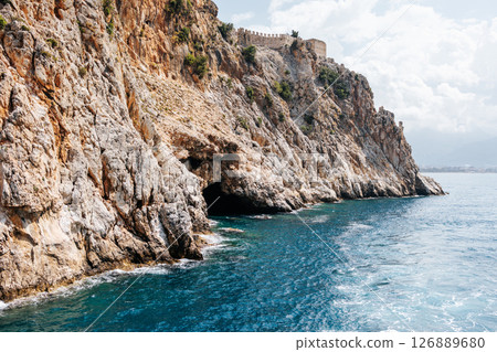 Sea cave in a rocky shore with turquoise transparent water. Picturesque natural landscape with rough texture of stones and deep entrance to the grotto. Ecotourism. Fascinating seascape. 126889680