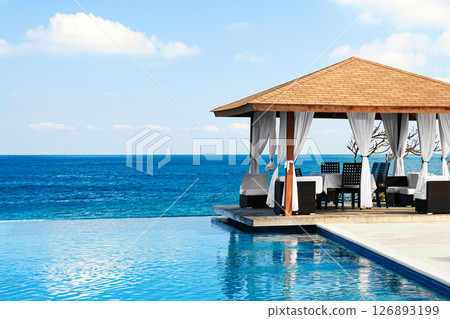 Beach with a infinite pool and a pavilion. The pavilion is covered with white curtains. The water is calm and the sky is clear 126893199