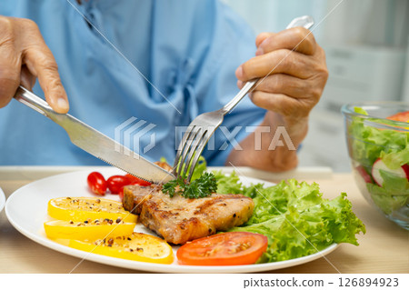 Asian senior woman patient eating pork chop stake and vegetable salad for healthy food in hospital. 126894923