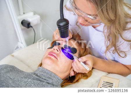 Eyelash extension procedure using ultraviolet lamp in modern beauty salon. Innovative lash enhancement technology with precise application technique. 126895136