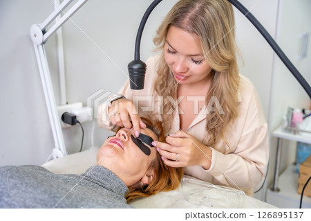 Beautician applying black eye patches before eyelash extension procedure with ultraviolet lamp. Innovative lash extension method in modern beauty salon 126895137