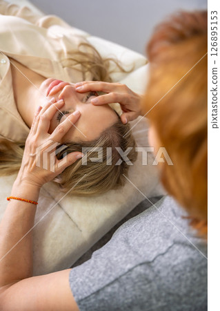 Female practitioner performs facial lifting technique on a young woman lying on a couch. Concept of energy healing, face therapy and alternative wellness practice from Access Bars. 126895153