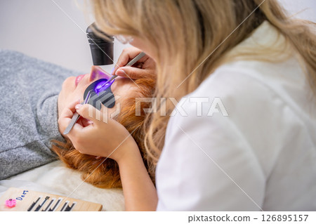 Eyelash extension procedure using ultraviolet lamp in modern beauty salon. Innovative lash enhancement technology with precise application technique. Eyelash extension procedure using ultraviolet lamp in modern beauty salon. Innovative lash enhancement technology with precise application technique. 126895157
