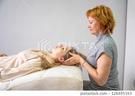 Female practitioner performs facial lifting technique on a young woman lying on a couch. Concept of energy healing, face therapy and alternative wellness practice from Access Bars. Female practitioner performs facial lifting technique on a young woman lying on a couch. Concept of energy healing, face therapy and alternative wellness practice from Access Bars. 126895163