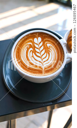 Cappuccino cup featuring intricate leaf latte art, resting on wooden surface, bathed in warm natural light filtering through glass window 126895467