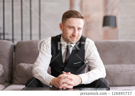 A man in business clothes sits at a table with folded arms in a modern interior with soft lighting. A focused look and professional style emphasize the atmosphere of confidence and professionalism. 126895737