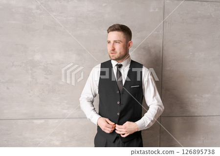 man in business clothes is buttoning up his vest, standing against a wall with a neutral background. His focused gaze and neat appearance emphasize the atmosphere of preparation and professionalism. man in business clothes is buttoning up his vest, standing against a wall with a neutral background. His focused gaze and neat appearance emphasize the atmosphere of preparation and professionalism. 126895738