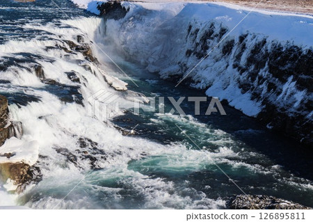 horizontal photo took in Gullfoss Falls, Hvita river, Golden Circle, Reykjavik, Iceland. A waterfall cascading over rocky cliffs into a river below with snow covered areas and a clear blue sky 126895811
