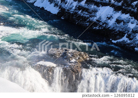 horizontal photo took in Gullfoss Falls, Hvita river, Golden Circle, Reykjavik, Iceland. A waterfall cascading over rocky cliffs into a river below with snow covered areas and a clear blue sky 126895812