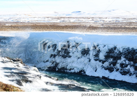 horizontal photo took in Gullfoss Falls, Hvita river, Golden Circle, Reykjavik, Iceland. A waterfall cascading over rocky cliffs into a river below with snow covered areas and a clear blue sky horizontal photo took in Gullfoss Falls, Hvita river, Golden Circle, Reykjavik, Iceland. A waterfall cascading over rocky cliffs into a river below with snow covered areas and a clear blue sky 126895820