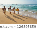 Four women walk on sandy beach holding surfboards. Female surfers silhouettes cast long shadows on sand. Ocean waves break gently nearby. Clear sky and horizon. Beach vibe with water sport activity. 126895859