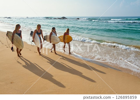 Four women walk on sandy beach holding surfboards. Female surfers silhouettes cast long shadows on sand. Ocean waves break gently nearby. Clear sky and horizon. Beach vibe with water sport activity. Four women walk on sandy beach holding surfboards. Female surfers silhouettes cast long shadows on sand. Ocean waves break gently nearby. Clear sky and horizon. Beach vibe with water sport activity. 126895859