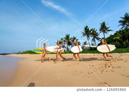 Four diverse female surfers walk with surfboards on tropical beach. Friends enjoy sunny day, palm trees. Women athletes ready for ocean waves. Active lifestyle, summer adventure. Four diverse female surfers walk with surfboards on tropical beach. Friends enjoy sunny day, palm trees. Women athletes ready for ocean waves. Active lifestyle, summer adventure. 126895860