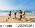 Four women holding surfboards run on tropical beach towards ocean waves. Bikini sport lovers enjoy surfing adventure, explore seaside holiday spot. Friends bond in surf trip, summer travel fun. 126895861
