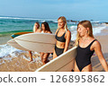 Women in swimsuits carry surfboards on sunny beach. Surfing tour. Friends ready for adventure on ocean waves. Enjoying travel, active lifestyle, tropical day. Smiling faces share joy. 126895862