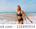 Blonde woman in black bikini with surfboard on sandy beach. Smiling surfer stands by ocean waves. Summer water sport activity, surfing adventure. Holiday at tropical sea coast. Sunny day. 126895914
