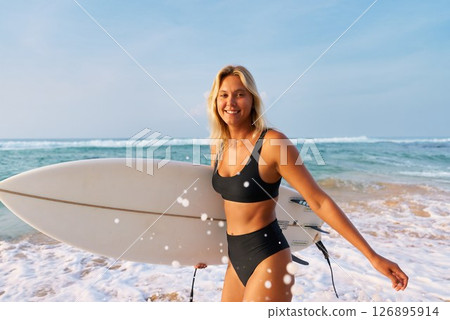 Blonde woman in black bikini with surfboard on sandy beach. Smiling surfer stands by ocean waves. Summer water sport activity, surfing adventure. Holiday at tropical sea coast. Sunny day. Blonde woman in black bikini with surfboard on sandy beach. Smiling surfer stands by ocean waves. Summer water sport activity, surfing adventure. Holiday at tropical sea coast. Sunny day. 126895914