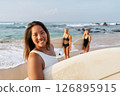 Smiling woman on sandy beach holds surfboard. Friends in swimsuits behind prepare for surfing. Ocean waves and sunny weather. Summer fun, water sports, beach vibes. 126895915