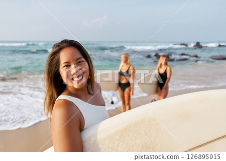 Smiling woman on sandy beach holds surfboard. Friends in swimsuits behind prepare for surfing. Ocean waves and sunny weather. Summer fun, water sports, beach vibes. Smiling woman on sandy beach holds surfboard. Friends in swimsuits behind prepare for surfing. Ocean waves and sunny weather. Summer fun, water sports, beach vibes. 126895915