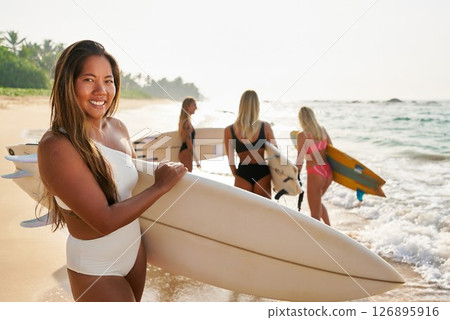 Group of young women with surfboards walk on sunny tropical beach. Surfers wear swimwear and prepare for ocean surfing. Friends enjoy water sport, adventure and travel at seaside holiday. Group of young women with surfboards walk on sunny tropical beach. Surfers wear swimwear and prepare for ocean surfing. Friends enjoy water sport, adventure and travel at seaside holiday. 126895916
