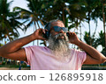 Elderly man with beard wearing headphones stands relaxed. Listening to ethnic tropical music under palm trees. Outdoor summer vibe. Vacation mood. Island vibes. 126895918