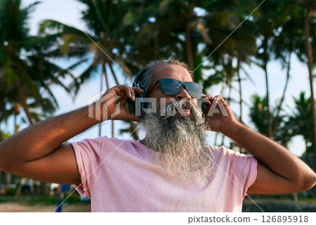 Elderly man with beard wearing headphones stands relaxed. Listening to ethnic tropical music under palm trees. Outdoor summer vibe. Vacation mood. Island vibes. 126895918
