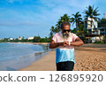 Senior man uses smart watch on tropical beach. Bearded grandpa with earbuds checks fitness tracker. Modern tech, active lifestyle, ocean background. Elderly enjoys digital innovation, relaxation. 126895920