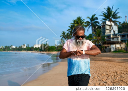 Senior man uses smart watch on tropical beach. Bearded grandpa with earbuds checks fitness tracker. Modern tech, active lifestyle, ocean background. Elderly enjoys digital innovation, relaxation. Senior man uses smart watch on tropical beach. Bearded grandpa with earbuds checks fitness tracker. Modern tech, active lifestyle, ocean background. Elderly enjoys digital innovation, relaxation. 126895920