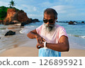 Senior ethnic man with grey beard on beach uses smart watch and earbuds for fitness. Elderly male enjoys tech for training, wellbeing. Old man in modern scene healthy lifestyle. 126895921
