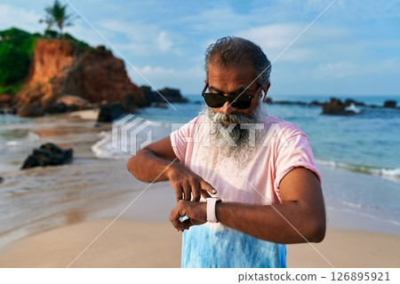 Senior ethnic man with grey beard on beach uses smart watch and earbuds for fitness. Elderly male enjoys tech for training, wellbeing. Old man in modern scene healthy lifestyle. Senior ethnic man with grey beard on beach uses smart watch and earbuds for fitness. Elderly male enjoys tech for training, wellbeing. Old man in modern scene healthy lifestyle. 126895921