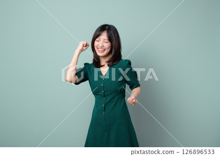 Southeast asian woman in green dress smiling with eyes closed and fist raised in joyful celebration against plain background, expressing happiness and success 126896533