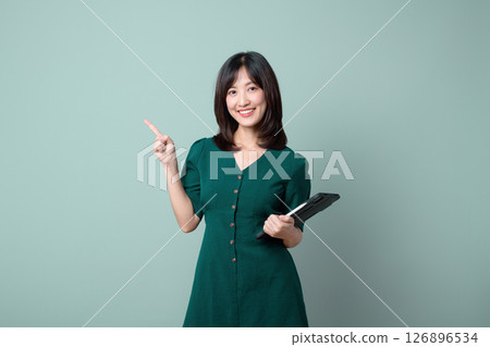 Southeast asian woman smiling while holding tablet and pointing upward, standing against plain background, representing internet technology and positive emotion Southeast asian woman smiling while holding tablet and pointing upward, standing against plain background, representing internet technology and positive emotion 126896534