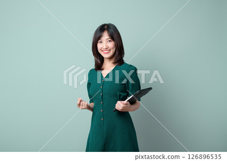 Happy Asian woman smiling and holding tablet, wearing green dress, standing isolated on light green background 126896535
