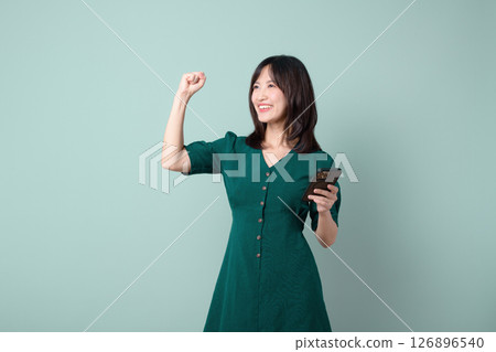 Asian woman in green dress celebrates good news on mobile phone, smiling and raising fist against green background Asian woman in green dress celebrates good news on mobile phone, smiling and raising fist against green background 126896540