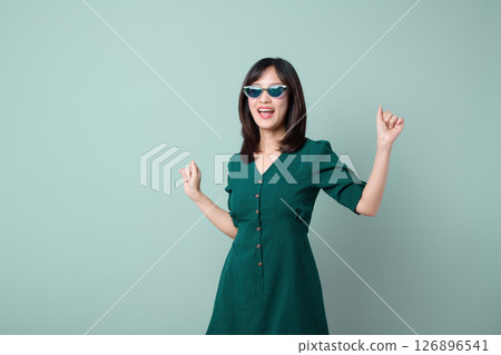 Southeast Asian woman in green dress wearing sunglasses stands against pastel background expressing happiness and confidence with joyful pose and cheerful smile Southeast Asian woman in green dress wearing sunglasses stands against pastel background expressing happiness and confidence with joyful pose and cheerful smile 126896541