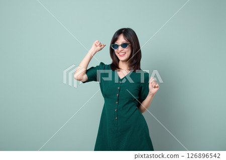 Southeast Asian woman wearing sunglasses and green dress smiling and dancing joyfully against plain light green background, expressing happiness and confidence Southeast Asian woman wearing sunglasses and green dress smiling and dancing joyfully against plain light green background, expressing happiness and confidence 126896542