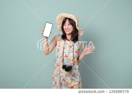 Cheerful southeast asian woman in floral dress and straw hat holding smartphone and camera, smiling against pastel green background, expressing happiness and excitement Cheerful southeast asian woman in floral dress and straw hat holding smartphone and camera, smiling against pastel green background, expressing happiness and excitement 126896578