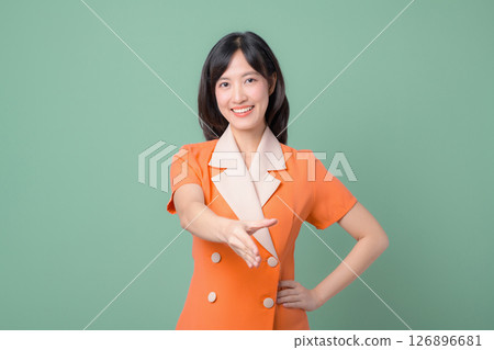 Southeast asian woman in orange suit smiling and extending hand for handshake against green background friendly gesture Southeast asian woman in orange suit smiling and extending hand for handshake against green background friendly gesture 126896681
